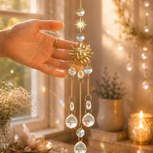May include: A gold-toned sun-shaped suncatcher featuring a central face design with radiating rays, suspended from a delicate metal chain. The ornament includes multiple clear, faceted glass crystals in various shapes that hang from the sun.