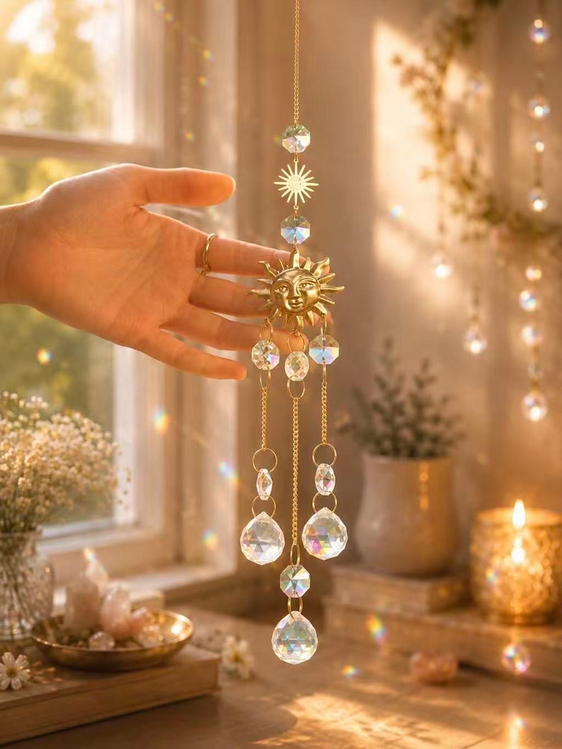 May include: A gold-toned sun-shaped suncatcher featuring a central face design with radiating rays, suspended from a delicate metal chain. The ornament includes multiple clear, faceted glass crystals in various shapes that hang from the sun.