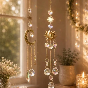 May include: Two hanging suncatchers suspended by gold-toned chains. One features a crescent moon charm, the other a sun face charm. Both are adorned with clear, multifaceted crystal beads and prisms that reflect light, creating a decorative display.