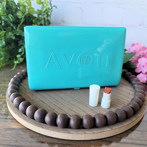 Puede incluir: Un estuche compacto Avon turquesa con el nombre de la marca en relieve. Un tubo de lápiz labial blanco y un lápiz labial con un tono rojo-naranja están junto al estuche. Los artículos están sobre una bandeja de madera con cuentas marrones.