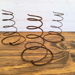 May include: Three antique, rusty metal springs with a spiral design. The springs are brown and sit on a wooden surface. The background includes a white brick wall.