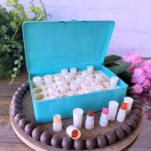May include: A rectangular turquoise storage box with a hinged lid, containing multiple white cylindrical lipstick tubes arranged in a grid. Several individual lipstick tubes with white caps and various colored tips are placed in front of the box.