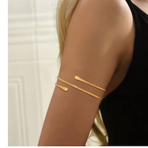 May include: Gold-toned arm cuff jewelry. The cuff features two parallel bands that wrap around the upper arm. The ends of the bands have a flattened, elongated design. The jewelry is worn on a tanned arm, with a black top in the background.