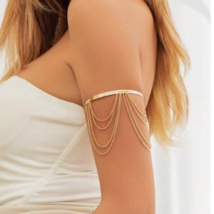 May include: Gold-coloured arm cuff featuring multiple dangling chain strands. The cuff is a simple band that sits above the elbow, with several delicate chains cascading down the arm. The jewellery is a stylish accessory.
