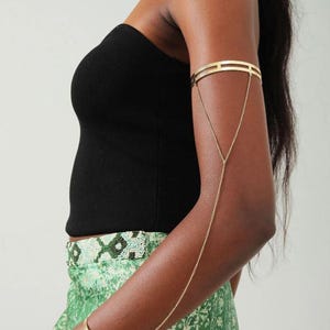 May include: Gold arm cuff and bracelet linked by a fine chain. The arm cuff has a geometric pattern, and the bracelet is a simple, curved design. The jewellery is worn on an arm, paired with a black top and green skirt.