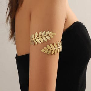 May include: Gold-colored arm cuffs with a leaf design. The cuffs are worn on the upper arm and are made of a shiny metal. The arm cuffs are a decorative accessory.
