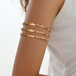 May include: Three gold arm cuffs with a textured, wavy design. The arm cuffs are worn on the upper arm. The jewellery is a bright gold colour and appears to be made of metal. The background is a white top.
