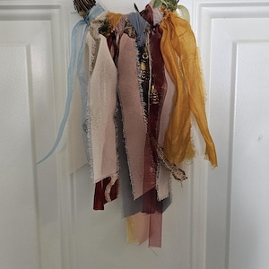May include: A circular wreath made of brown twisted twigs and green leaves, featuring a collection of hanging fabric strips in shades of mustard yellow, burgundy, dusty rose, light blue, and cream with frayed edges and varying textures.