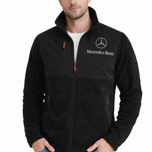 May include: Black fleece jacket with a full zipper, a stand-up collar, and red zipper pulls. The Mercedes-Benz logo is embroidered on the chest in white. The jacket has two front pockets and a chest pocket.