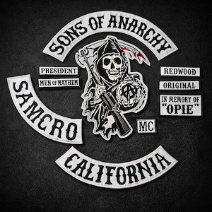 Può includere: Un insieme di toppe ricamate in bianco e nero. La toppa centrale raffigura una figura della morte con una pistola. Altre toppe includono "SONS OF ANARCHY", "SAMCRO", "CALIFORNIA", "PRESIDENT", "MEN OF MAYHEM", "REDWOOD", "ORIGINAL", "IN MEMORY OF 'OPIE'" e "MC".