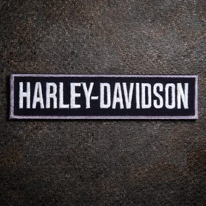 Op de afbeelding: Een rechthoekige zwarte patch met de geborduurde tekst "HARLEY-DAVIDSON" in het wit. De patch heeft een witte rand en is geplaatst tegen een getextureerde, donkerbruine achtergrond.