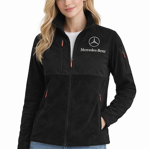 May include: Black fleece jacket with a full zipper, featuring the Mercedes-Benz logo in white on the chest. The jacket has a high collar, multiple pockets with orange zippers, and a soft, textured appearance. A comfortable outerwear option.