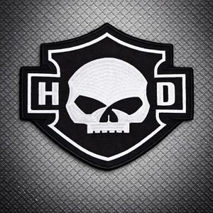 May include: A black and white patch with a skull design inside a shield shape. The letters "H" and "D" flank the skull. The patch has a white border and is likely an emblem or logo, possibly for a motorcycle club.
