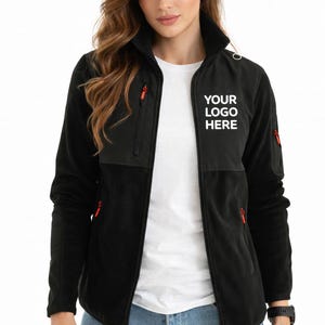 May include: Black fleece jacket with red zipper accents and the text "YOUR LOGO HERE" in white. The jacket has a high collar and two front pockets. The model is wearing a white shirt and blue jeans.