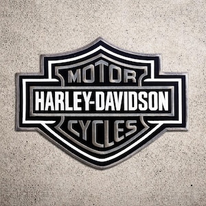 以下が含まれることがあります： ハーレーダビッドソンのロゴのクローズアップ。白文字で「MOTOR HARLEY-DAVIDSON CYCLES」と書かれた黒とシルバーのエンブレム。ロゴは、テクスチャ加工された明るい灰色の背景に配置されています。