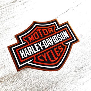 Puede incluir: Un parche de Harley-Davidson con el logotipo de la marca. El parche tiene forma de escudo con un borde marrón, detalles en blanco y negro y las palabras "MOTOR HARLEY-DAVIDSON CYCLES" en naranja y blanco.