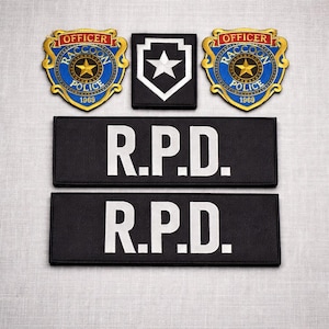 Può includere: Una collezione di toppe e nastri con nome ricamati. Due toppe presentano un disegno a scudo blu e oro con la scritta "OFFICER RACCOON POLICE 1969". Una stella bianca è centrata su una toppa a scudo in bianco e nero. Due nastri con nome neri leggono "R.P.D." in bianco.