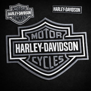 May include: A collection of Harley-Davidson logos in silver and black. The logos include the iconic shield shape with the words "MOTOR HARLEY-DAVIDSON CYCLES" and a rectangular logo with the words "HARLEY-DAVIDSON".