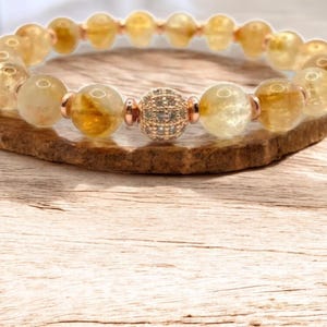 May include: A beaded bracelet featuring golden-yellow citrine gemstones and a central bead with a sparkling, silver-colored design. The bracelet is strung on a rose-gold colored chain and is displayed on a wooden surface.
