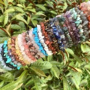 May include: A collection of gemstone chip bracelets in various colors, including turquoise, pink, and purple. The bracelets are stacked on top of each other, showcasing a variety of natural stone textures and hues. The background features green foliage.