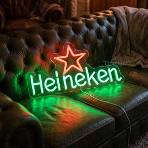 May include: A neon sign with the word "Heineken" in bright green, and a red star logo. The sign is placed on a dark green leather couch. The sign is lit up, creating a vibrant and eye-catching display.