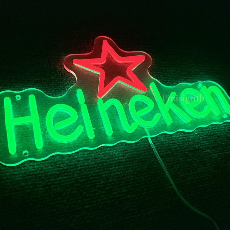 May include: A neon sign with the word "Heineken" in bright green, with a red star above the text. The sign is on a clear acrylic backing and has a power cord. The background is dark.