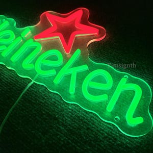 May include: A neon sign with the word "Heineken" in bright green, and a red star. The sign is made of clear acrylic and has a wavy outline. The sign is lit up and has a wire attached.