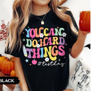 Puede incluir: Camiseta negra con la frase "YOU CAN DO HARD THINGS" en letras coloridas de estilo retro. El diseño incluye flores, estrellas, un lápiz y el hashtag "#testday".