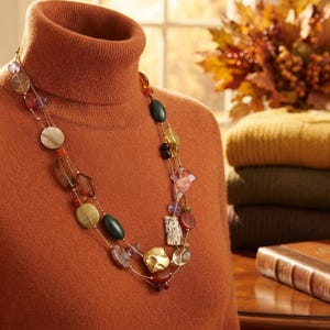 Op de afbeelding: Een meerstrengs ketting met een assortiment gepolijste kralen in diverse vormen, maten en kleuren, waaronder tinten groen, bruin, rood en goud, hangend aan dunne goudkleurige kettinkjes over een effen roestoranje coltrui.