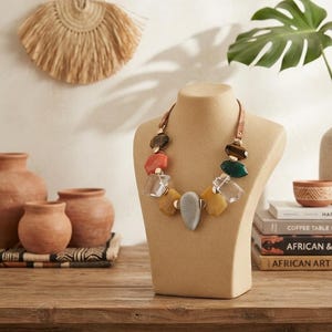 May include: A statement necklace featuring a variety of geometric beads in shades of brown, orange, clear, yellow, gray, and green, connected by a tan leather cord and displayed on a beige mannequin bust to showcase the multi-colored design.