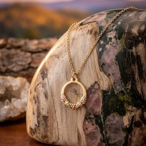 May include: A gold-toned circular pendant necklace featuring a small cluster of metallic leaves at the base. The pendant hangs from a delicate matching gold-toned chain, showcasing a polished finish with subtle color variations in the leaf detailing.