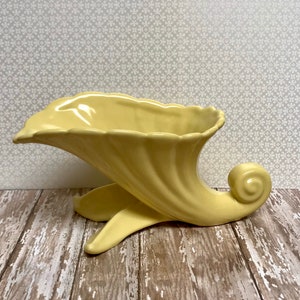 May include: A pale yellow ceramic cornucopia vase with a wide, flared opening and a curled horn. The vase is designed in a vintage style and is perfect for displaying flowers or other decorative items.