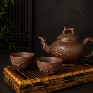 Puede incluir: Una tetera de arcilla marrón y dos tazas a juego con diseños en relieve de ramas de flor de ciruelo. La tetera tiene un asa y un remate de tapa esculpidos. El conjunto está dispuesto sobre una bandeja de bambú rectangular.