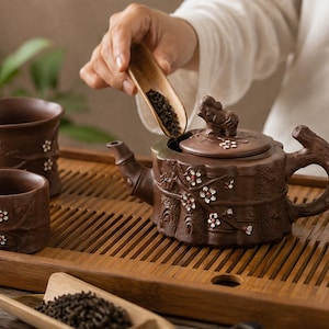 May include: A dark brown clay teapot and matching tea cups featuring embossed plum blossom branches with white and red petals. The teapot lid is topped with a small sculpted animal figure. The set is arranged on a slatted wooden tea tray.