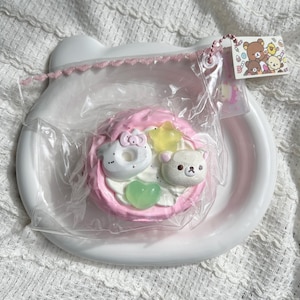 May include: A pink and white dessert-themed trinket, with a donut, bear, and star, on a white plate. The dessert is wrapped in clear plastic and has a tag with cartoon characters. The plate has a decorative edge.