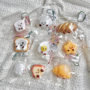 May include: A selection of food-themed keyrings, such as a slice of bread, a cloud, a croissant, a star, and an egg, each with a smiling face. The keyrings are packaged in clear plastic bags with attached key rings, set against a white, textured background.