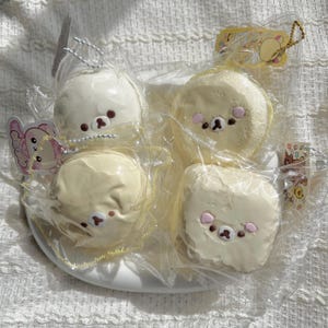 May include: Four plush bear-shaped toys in clear plastic packaging, arranged on a white plate. The bears are cream and light yellow with pink cheeks and brown facial features. Each toy has a small tag attached.