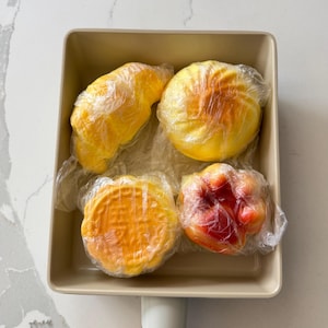 Op de afbeelding: Een beige vierkante pan met een houten handvat bevat vier verpakte voedingsmiddelen. De items zijn geel, oranje en rood en lijken gebak of bakwaren te zijn. De pan staat op een wit en grijs marmeren oppervlak.