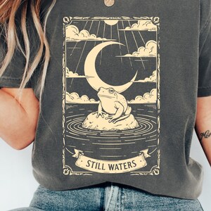 以下が含まれることがあります： 暗い灰色のTシャツ。クリーム色のイラストは、三日月の下の岩の上にいるカエルを描いています。デザインには雲、太陽光線、「Still Waters」という言葉がバナーに描かれています。 ゆったりとしたフィット感のシャツです。