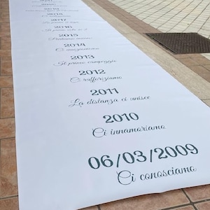Peut inclure: Une longue bannière en tissu blanc avec des dates et des phrases italiennes imprimées en gris foncé. La bannière énumère des dates importantes, commençant par "06/03/2009 Ci conosciamo" et se poursuivant avec des entrées annuelles. La bannière est posée sur une surface carrelée.