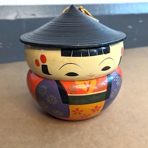 May include: A decorative, hand-painted Japanese Kokeshi doll-shaped container. The container is composed of two parts, a base and a lid. The base is painted with a colorful design of orange, purple, and yellow. The lid is black and conical, with a gold string.