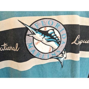 May include: A light blue and black striped shirt with the Florida Marlins logo embroidered on the front. The logo features a marlin leaping over a baseball, with the words "Florida Marlins" in a circular design.