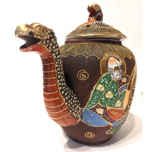 May include: A brown ceramic teapot with a dragon-shaped spout and handle. The teapot features a detailed painting of a seated figure in traditional attire, with gold and colorful accents. The lid is adorned with a small dragon figurine.