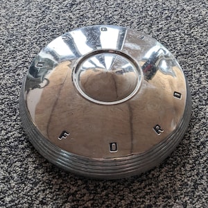 Puede incluir: Un tapacubos cromado brillante con la palabra "FORD" grabada en el borde. El diseño circular presenta un centro elevado y múltiples anillos concéntricos. La superficie refleja la luz.
