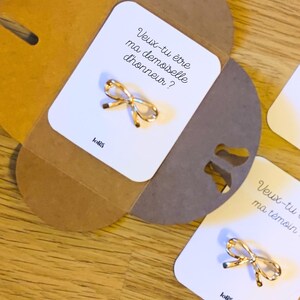 Peut inclure: Épinglette dorée en forme de nœud sur une carte blanche avec le texte "Veux-tu être ma demoiselle d'honneur ?" dans une enveloppe marron. Accessoire décoratif délicat.