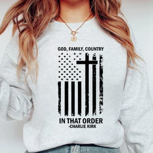 May include: A light grey sweatshirt with a distressed American flag design, a cross, and the text "GOD, FAMILY, COUNTRY IN THAT ORDER -CHARLIE KIRK" in black. The sweatshirt has a crew neck.