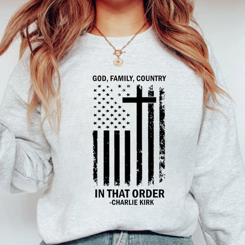 May include: A light grey sweatshirt with a distressed American flag design, a cross, and the text "GOD, FAMILY, COUNTRY IN THAT ORDER -CHARLIE KIRK" in black. The sweatshirt has a crew neck.