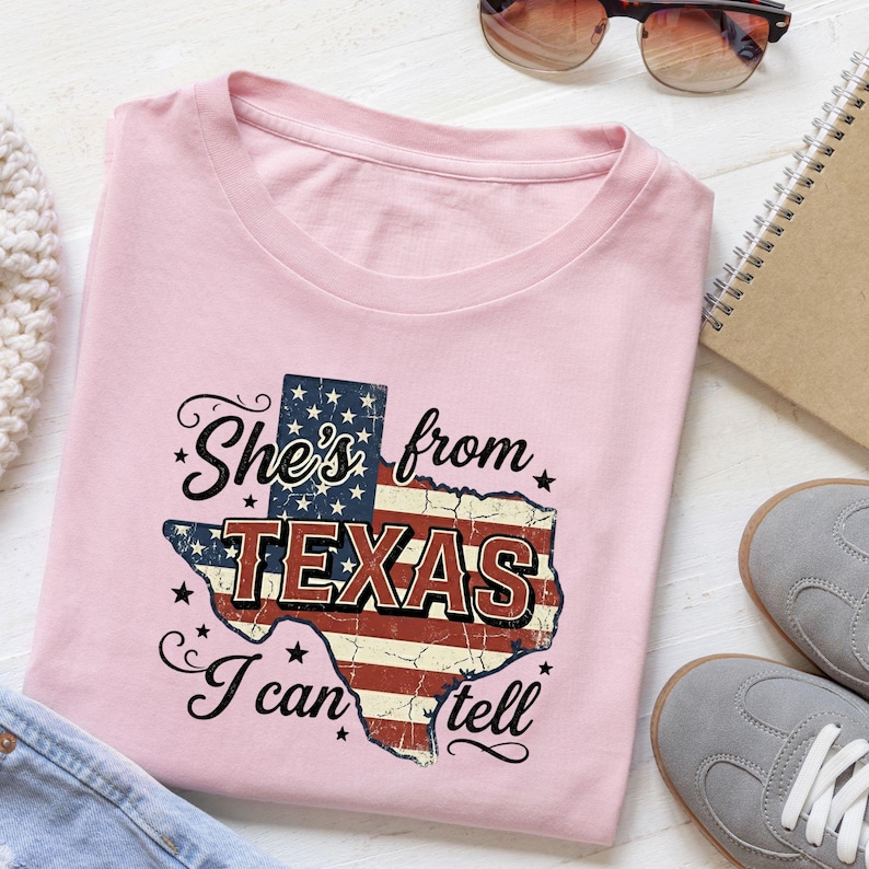 Pode incluir: Uma t-shirt rosa claro com um gr&aacute;fico do estado do Texas preenchido com um padr&atilde;o de bandeira americana desgastado. O texto "She's from TEXAS I can tell" est&aacute; impresso em letras pretas cursivas e de bloco sobre a forma do estado com estrelas.