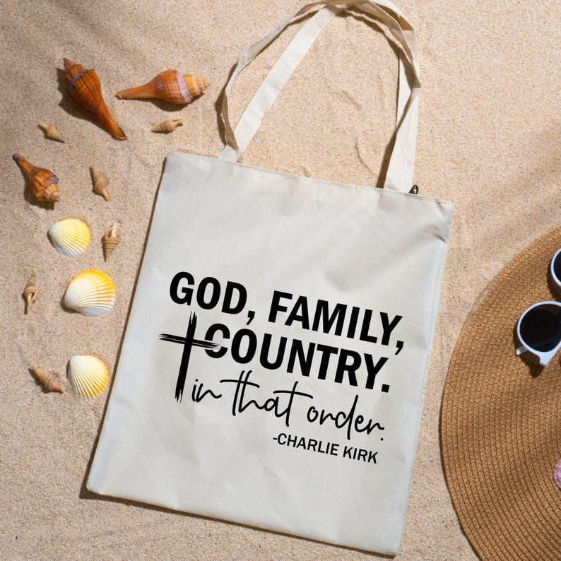 May include: A cream tote bag with the text "GOD, FAMILY, COUNTRY, in that order." in black. The bag has long handles and is on a sandy surface with seashells and a straw hat.