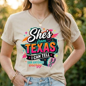Pode incluir: Uma t-shirt bege de manga curta com um gr&aacute;fico preto do estado do Texas. O texto diz She's from TEXAS I CAN TELL e SAN ANTONIO energy, com detalhes coloridos em rosa, laranja e turquesa, al&eacute;m de um pequeno log&oacute;tipo de basquetebol.